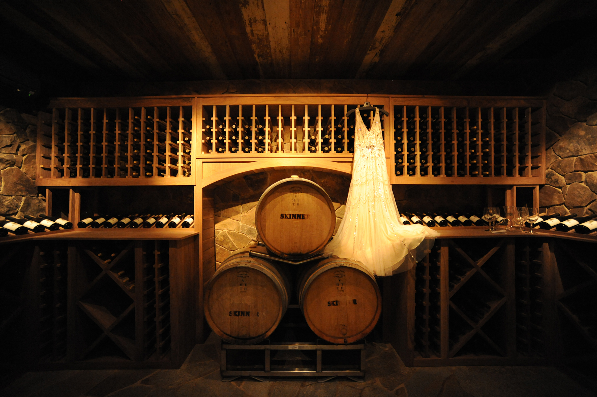 wine barrels and wine bottles in a cellar with the wedding dress hanging beside them