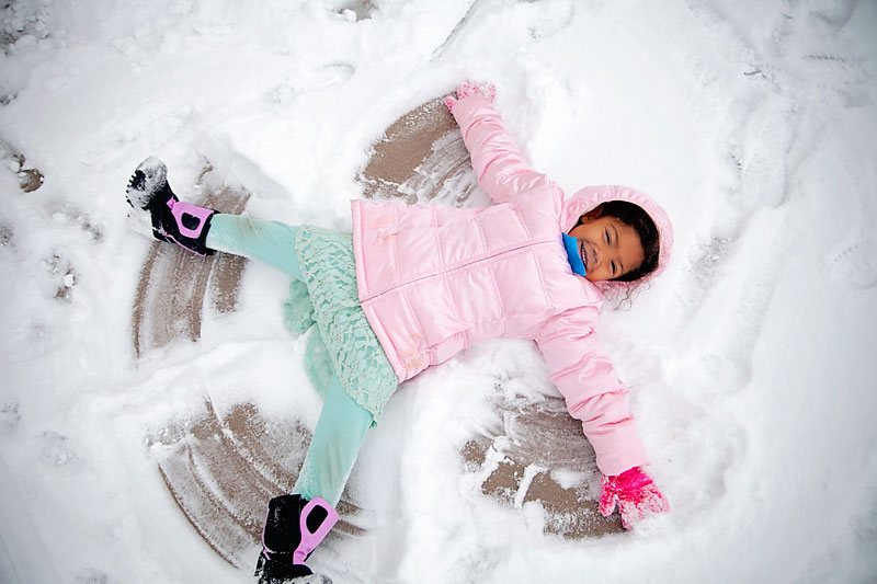 Young girl in pink parka, mint pants, pink snow boots, and pink gloves making a snow angel