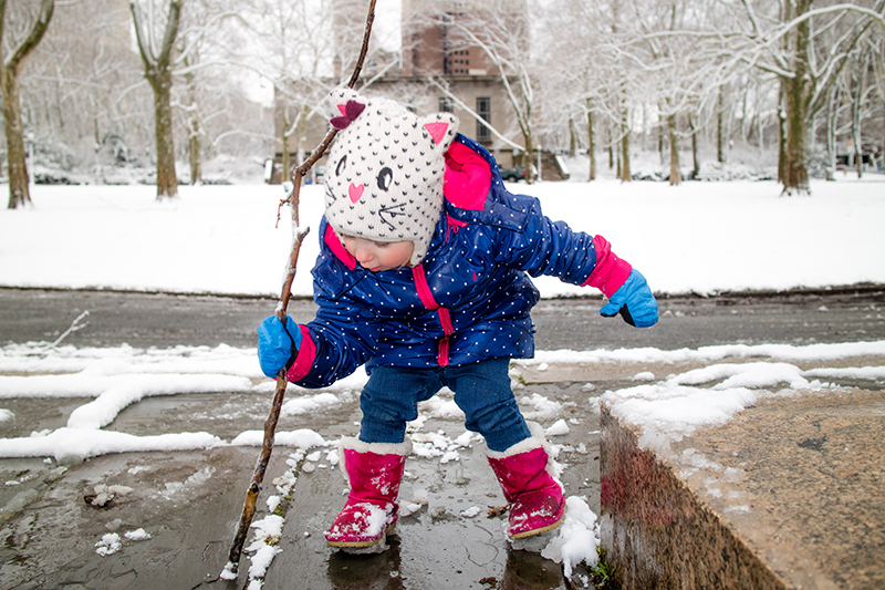 Young girl in blue polka dot parka and cat snow hat playing with a branch in the snow