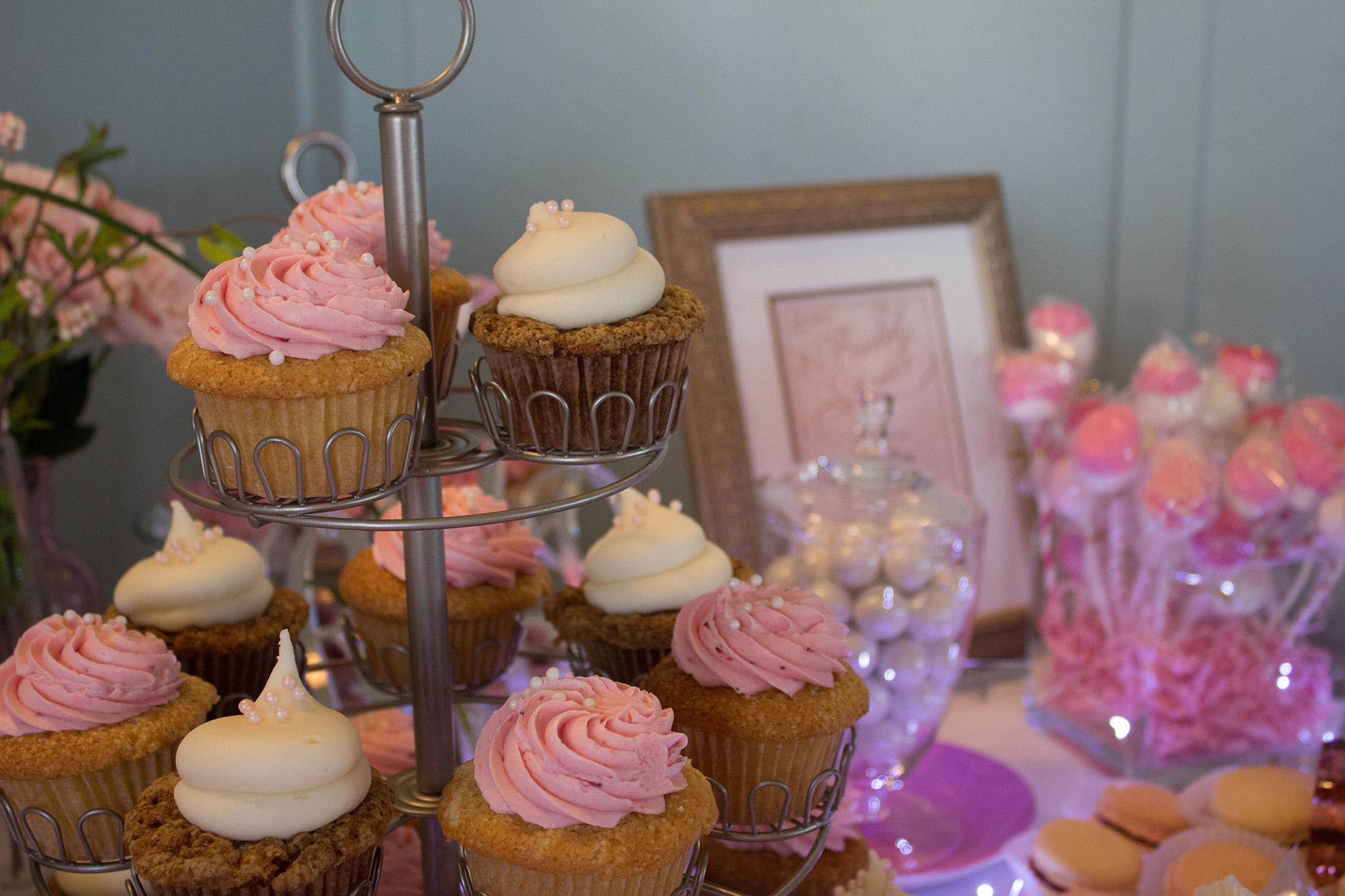three tiered cupcake stand with pink and white frosted cupcakes and other assorted pink treats in the background