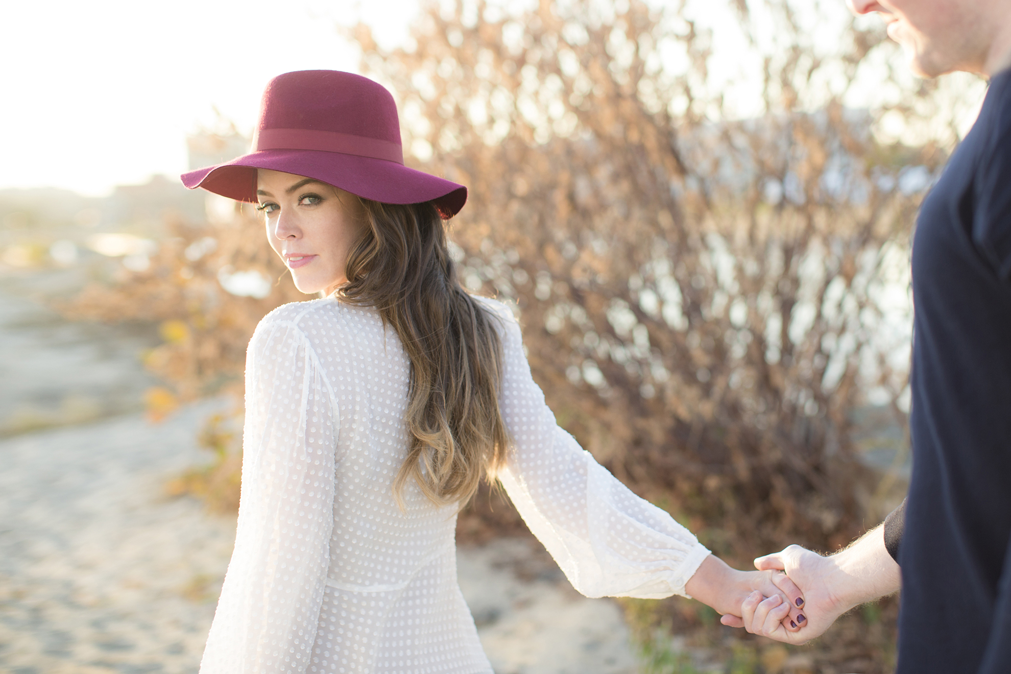 Image of a woman on the beach in a red brimmed hat and white blouse holding her partner's hand while looking back at the camera