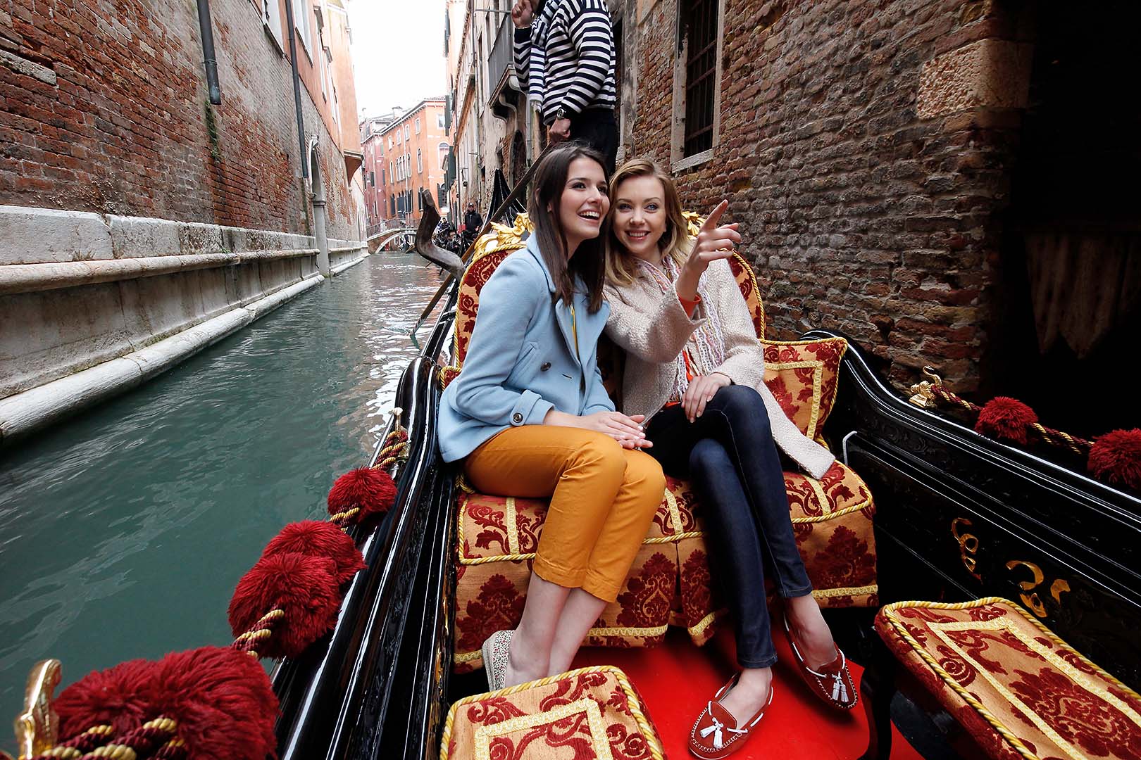 portrait of two women sitting on  red and yellow cusions in a gondola along a river of water between two brick buildings