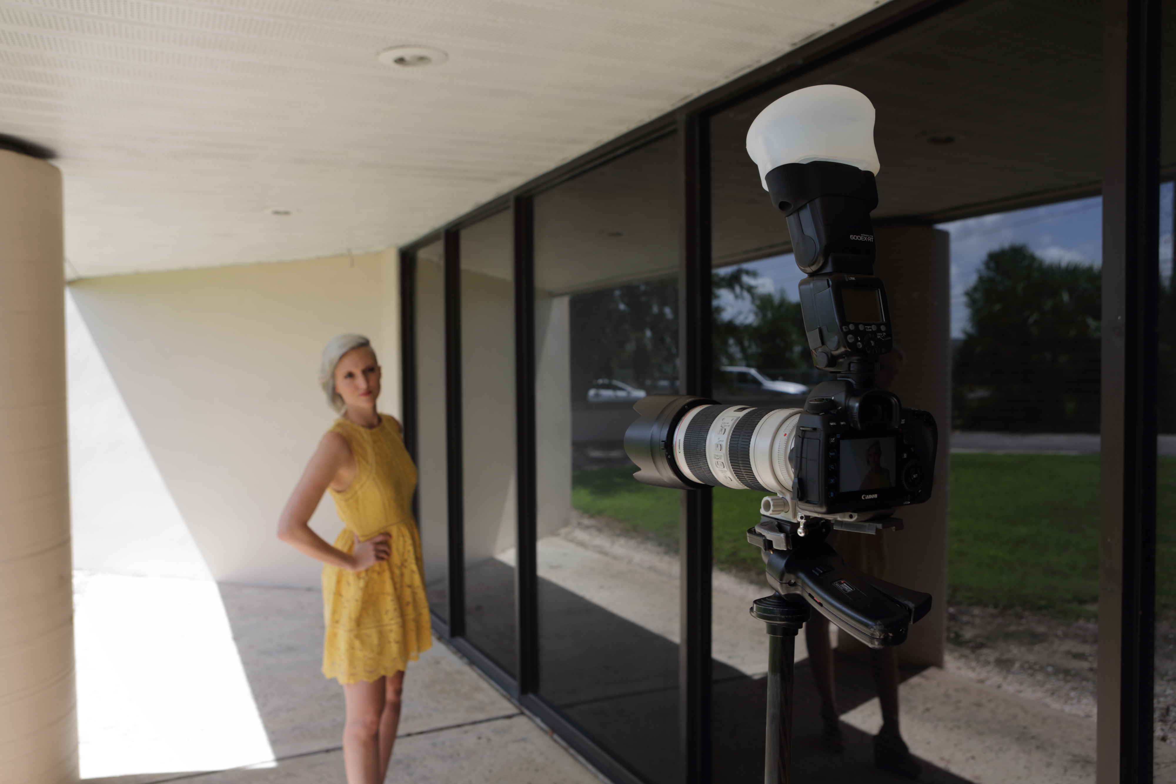 Blonde model standing in a yellow dress alongside a wall of windows with a camera and speedlite taking her photo