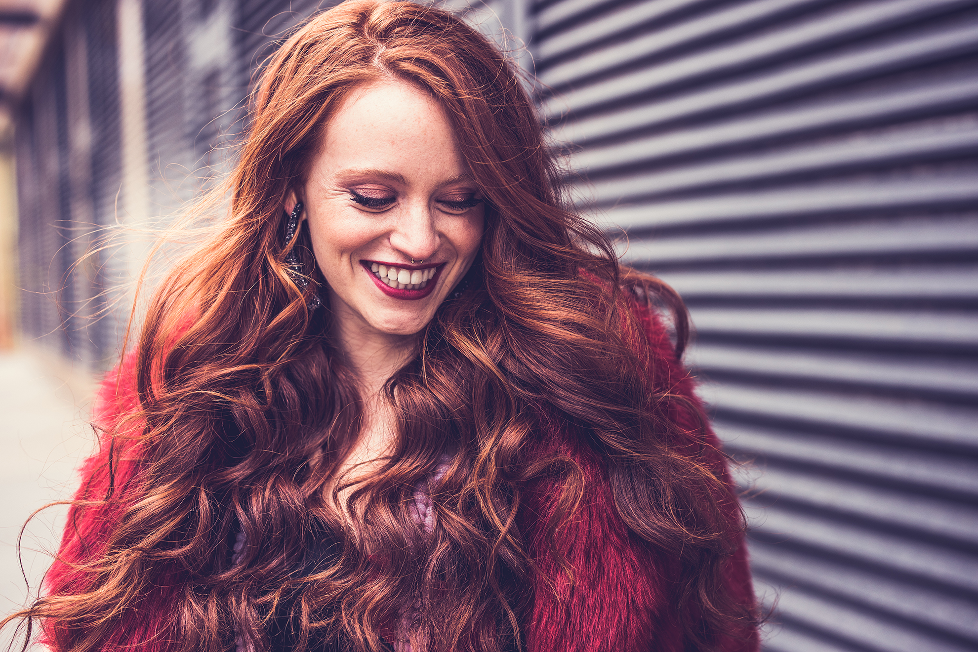 Red-haired model portrait smiling down at the ground with bright red lipstick and matching red fur coat