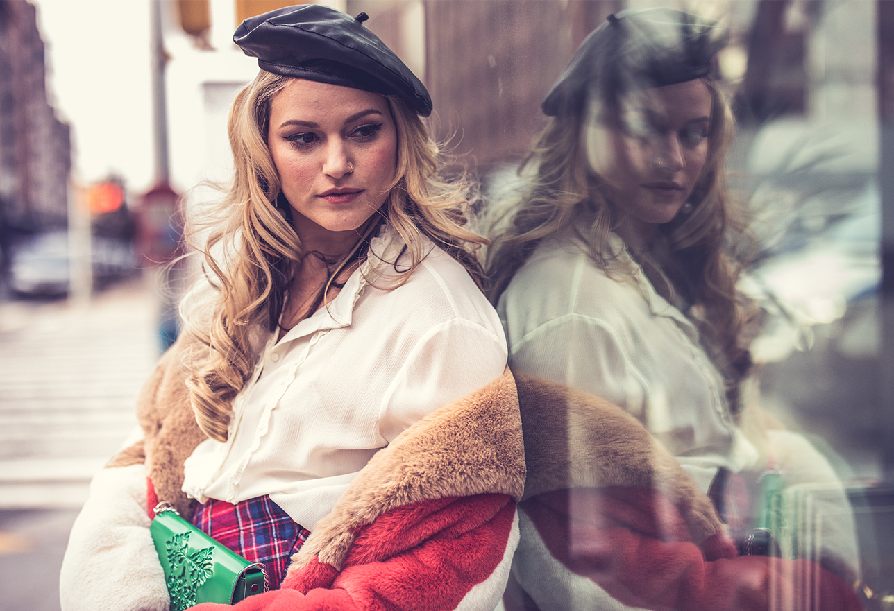Blonde model wearing a beret and holding an embossed green clutch looking through a shop window on a NYC sidewalk