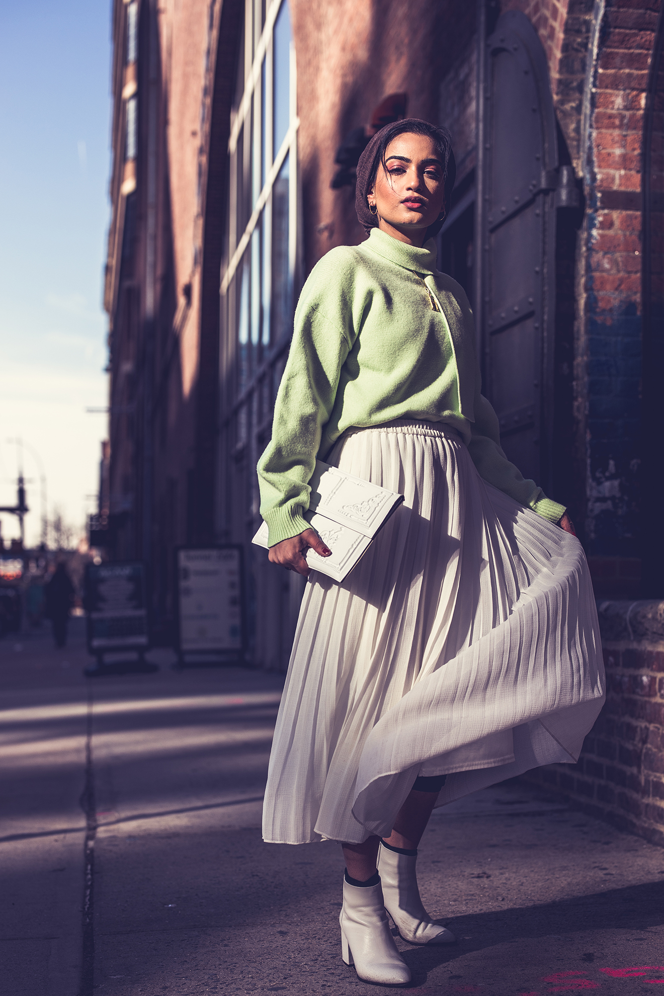 Dark haired model in a green cowel neck sweater and long white accordioned skirt posing on the sidewalk ear a large brick building