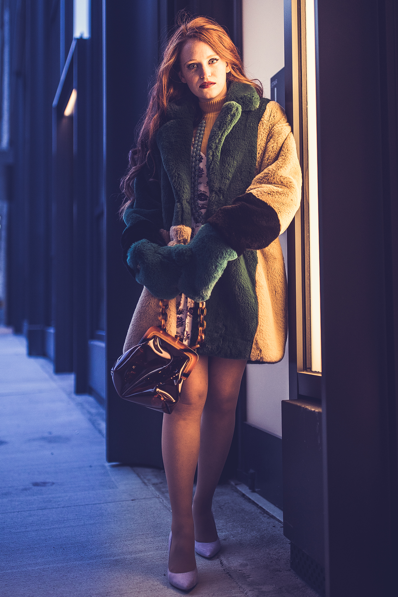 Nighttime shot of red-haired model in a green and beige fur jacket stanidng beside an illuminated shop window