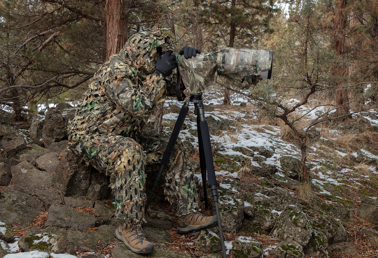 Image of photographer looking through camera that is perched on a tripod and completely covered in camoflauge