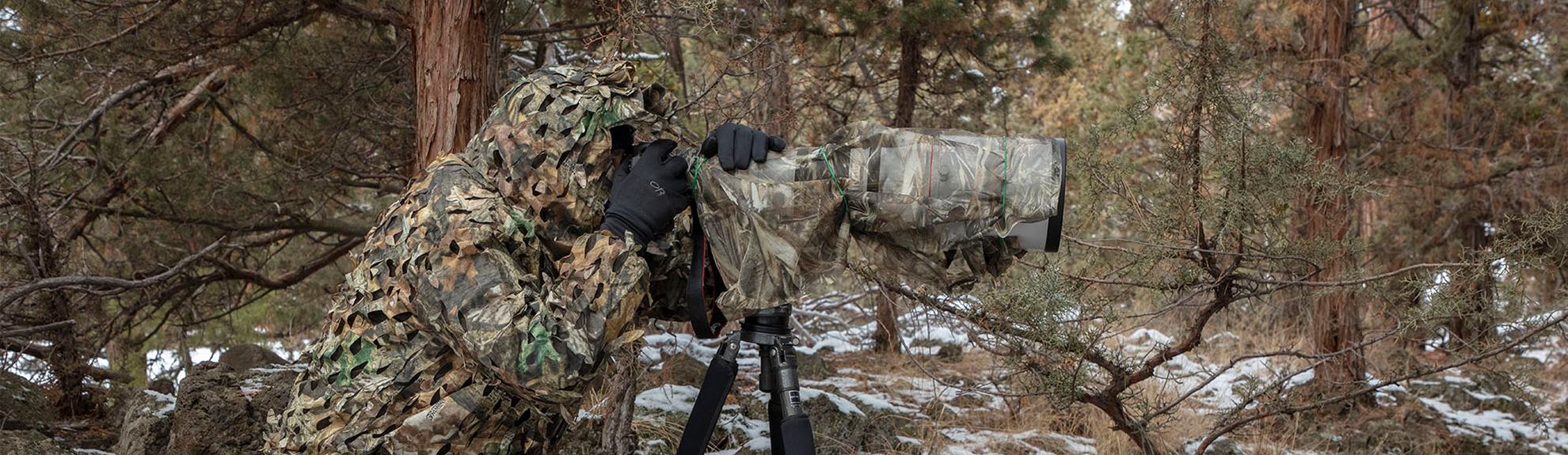 Image of photographer looking through camera that is perched on a tripod and completely covered in camoflauge