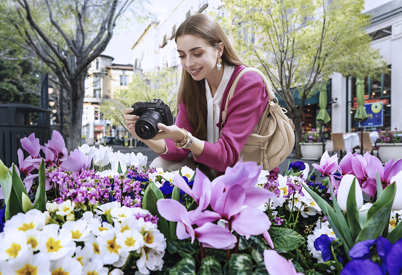 Woman kneeling in front of purple and white flowers wearing a pink cardigan, a beige purse, and holding her full-frame Canon camera