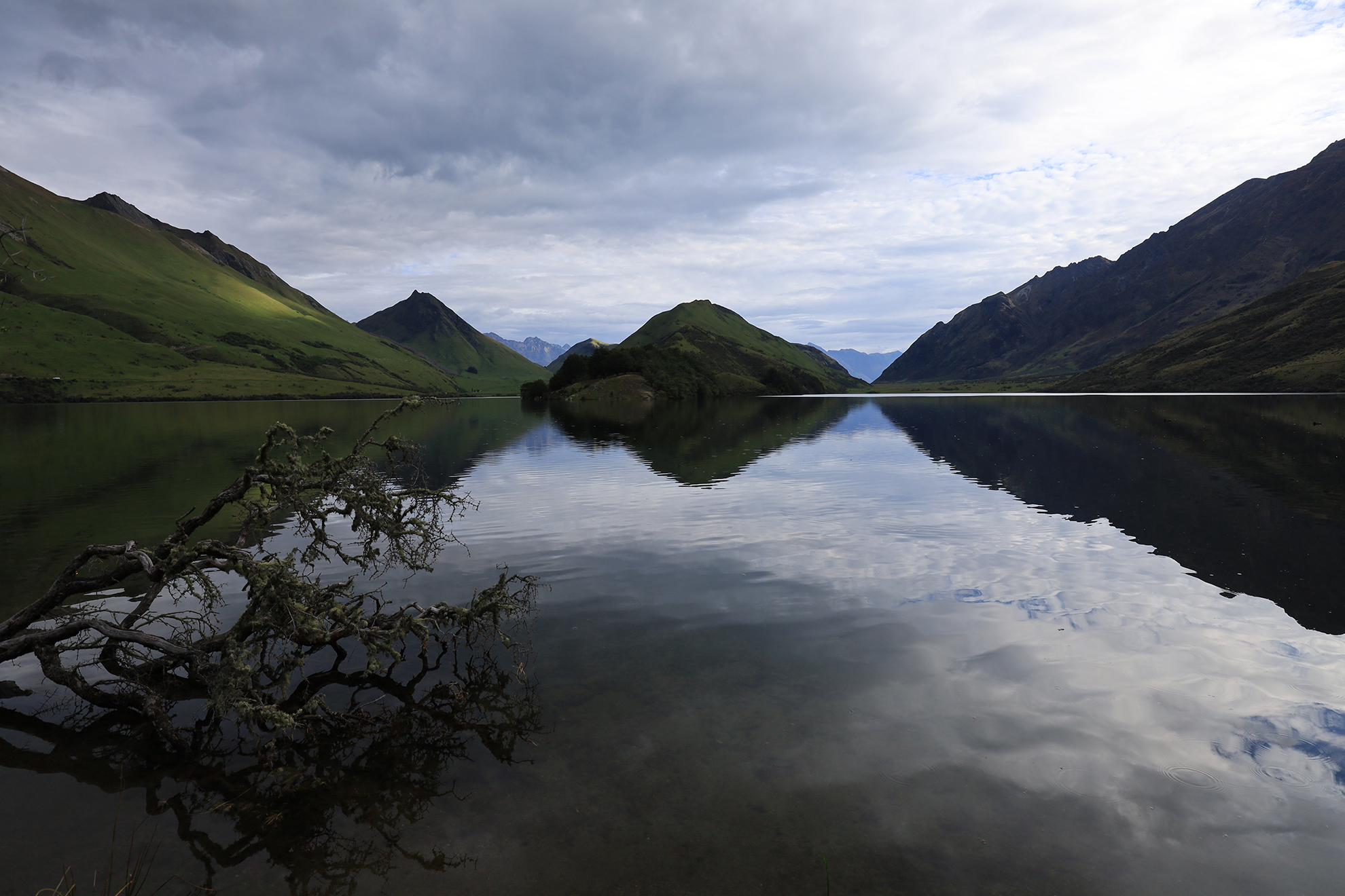 Wide-angle shot of a lake with green mountains in the backgroud and clouds reflecting in the water